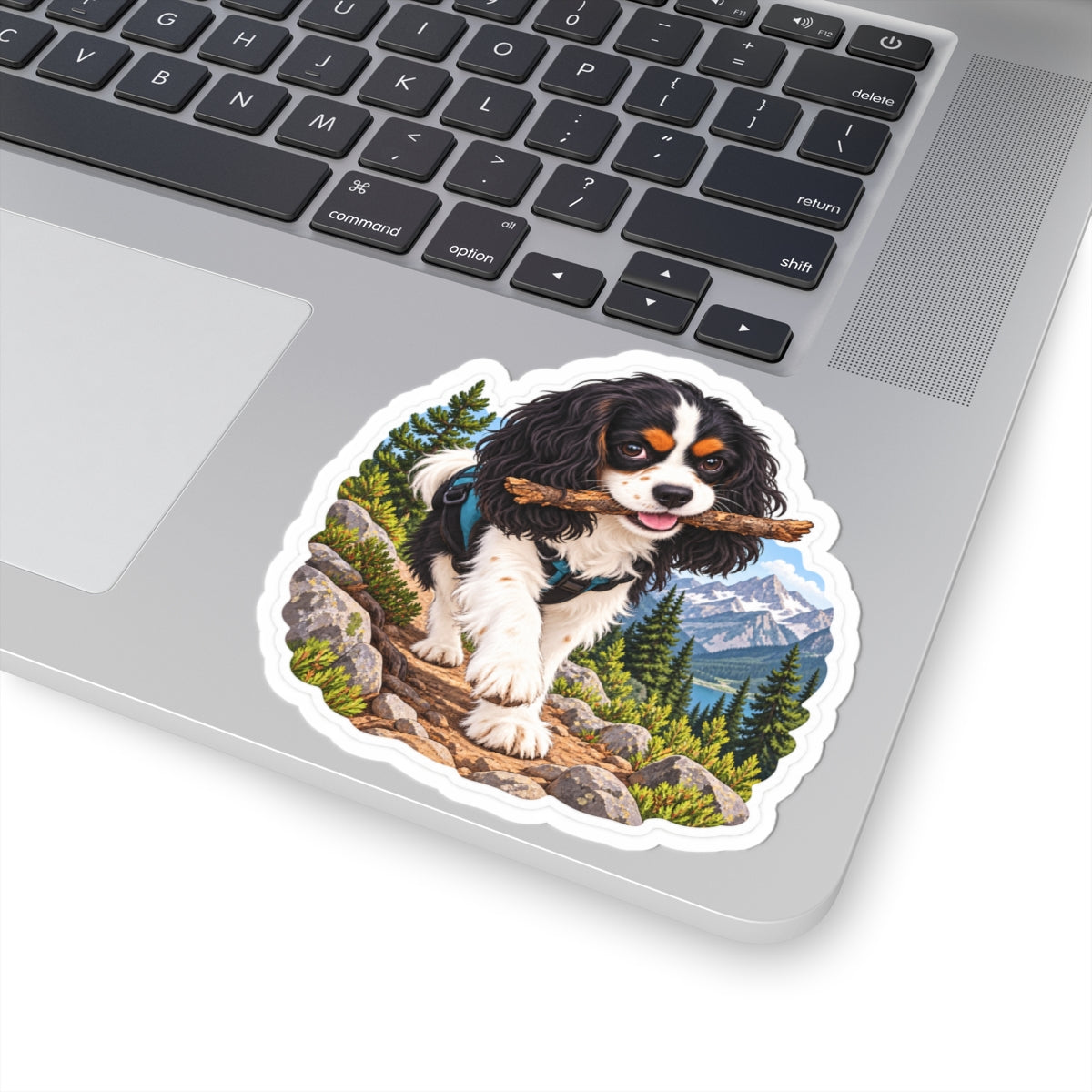 Product mockup of the Cavy Play Mode Trail Stick kiss-cut sticker: a tricolor Cavalier King Charles Spaniel carrying a stick along a mountain trail with pines and snowy peaks.